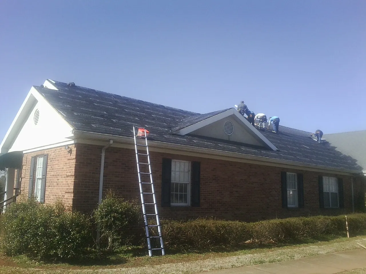 Crew tearing shingles off a brick ranch roof with a ladder set — Metal Roof Repair work by our crew in Mc Farlan