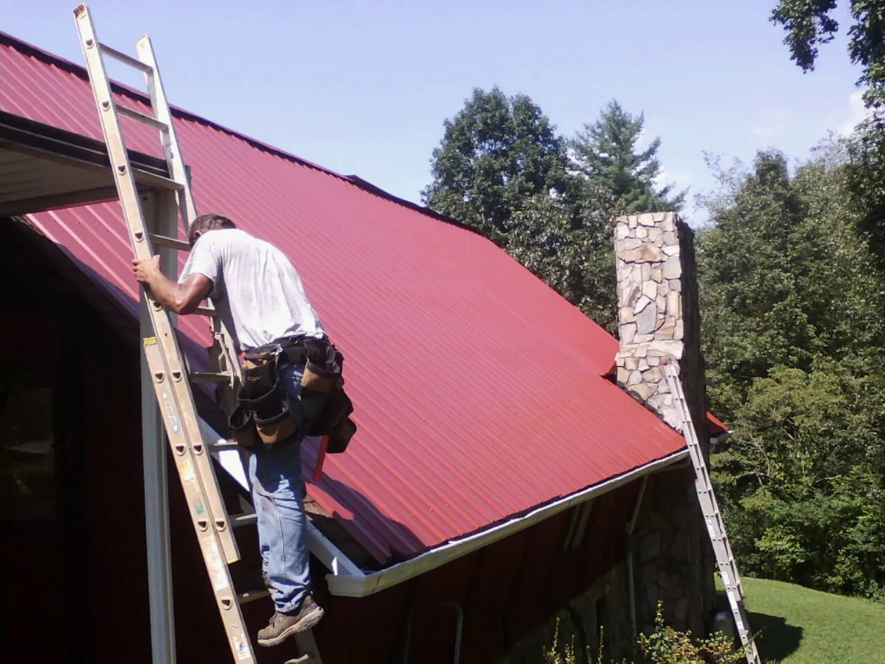 Roofer on a ladder beside a red metal roof and stone chimney — Soffit & Fascia Repair by Roofing Brothers in Mc Farlan